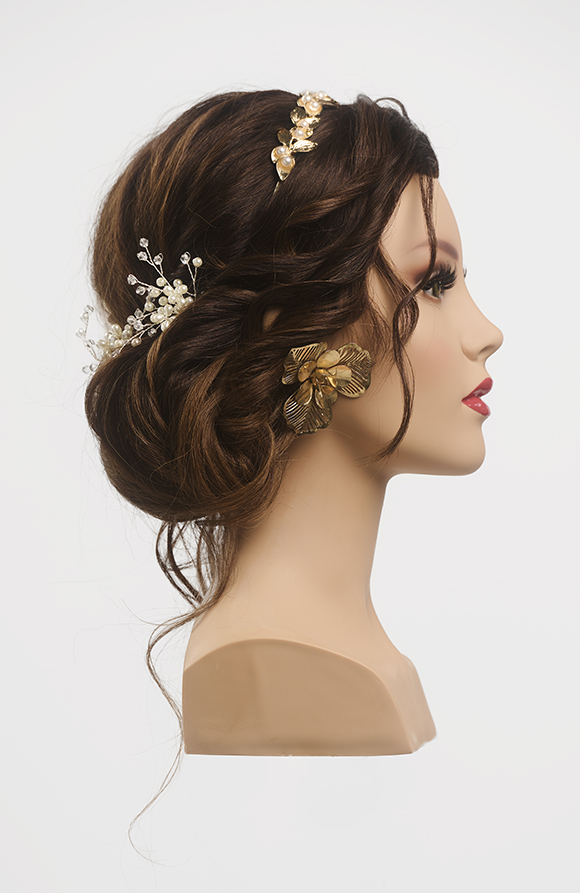 Brünetter, langhaariger Frisurenkopf im Profil, die Haare sind zu einem niedrigen Dutt gesteckt, am Oberkopf ist ein goldenes Stirnband zu sehen, der Kopf trägt große goldene Blumenohrstecker