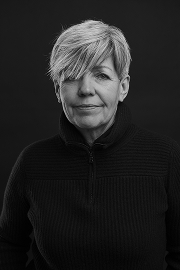 Black and white frontal portrait of a middle-aged female, short light hair covering her right eye, black sweater on black background; she smiles slightly into the camera; her name is Gabriele Bürger, Founder
