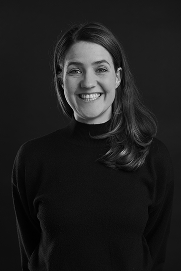 Black and white portrait, young female person with long dark hair, in black sweater against black background; she grins broadly into the camera. Adina Alt, CEO of L'IMAGE 
