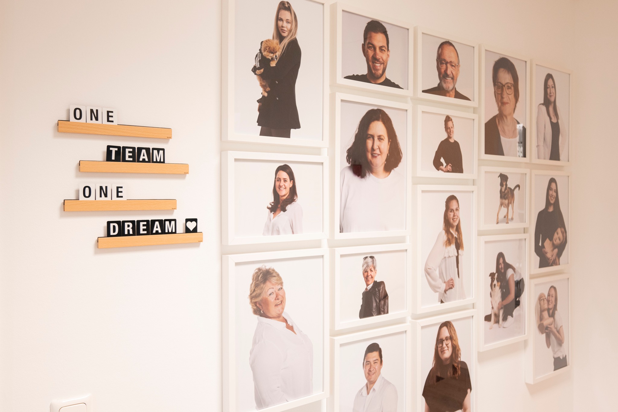 A white wall with several portraits of the L'IMAGE team in white picture frames. To the left of the picture frames hang four decorative wooden ledges, one word per ledge "ONE TEAM ONE DREAM". 