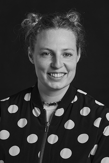 Schwarzweißes Portrait einer jungen weiblichen Person, die Haare zu Spacebuns geknotet, in einer schwarzen Jacke mit großen weißen Punkten; sie grinst breit in die Kamera; ihr Name ist Julia Siemons, Marketing