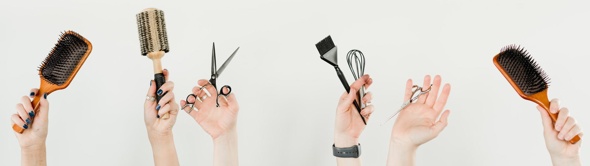 Six hands, visible from the wrist, hold different hairdressing utensils at the bottom of the picture, for example hairbrushes, haircutting scissors and dyeing brushes
