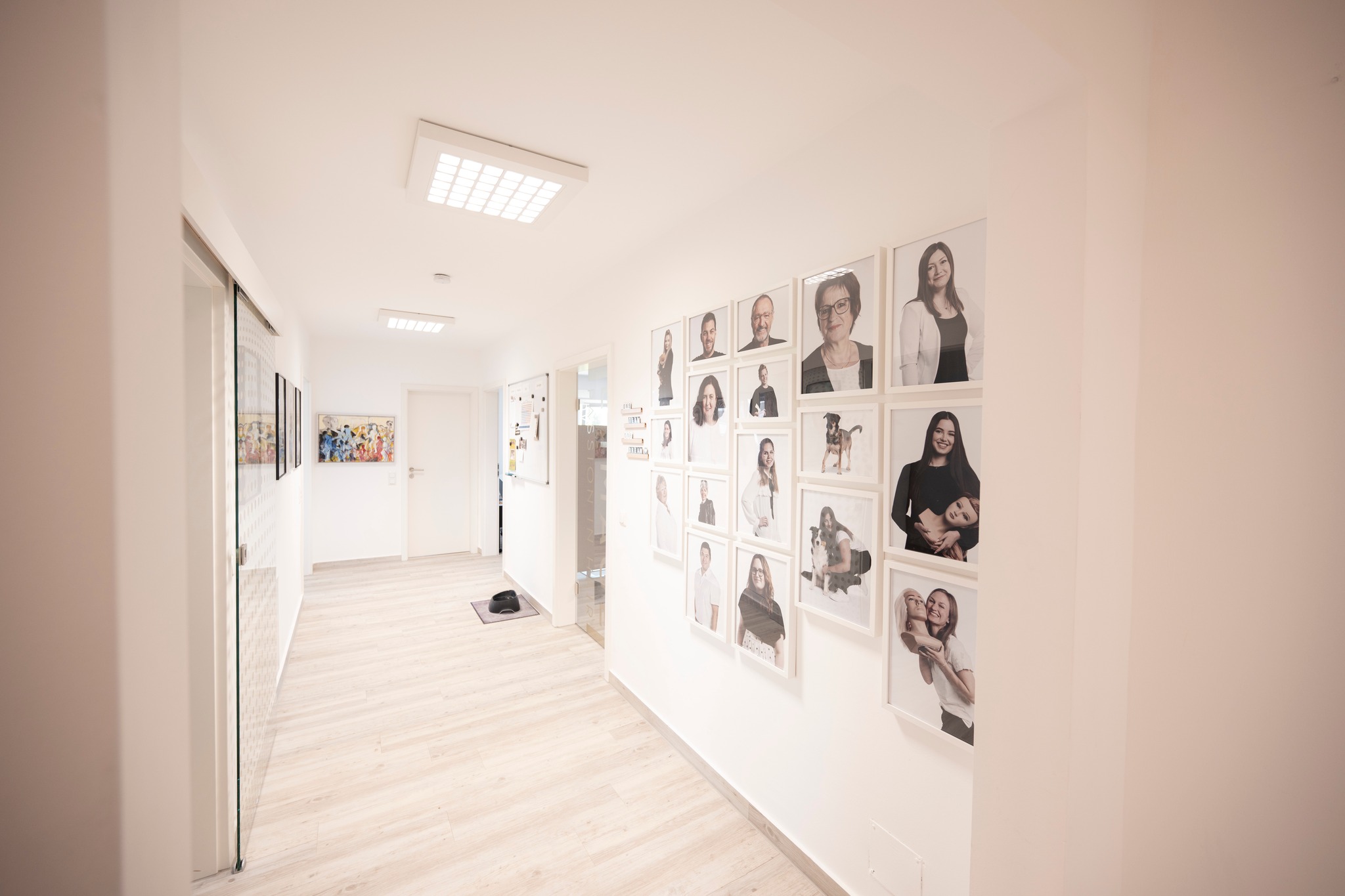 A long, brightly lit corridor, on the right side hang portraits of the team, behind them follow two doors; on the left side you can see a door, and at the end of the corridor also a white door